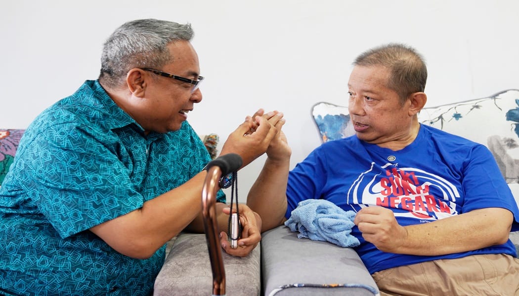 Two men on a couch clasping hands in a friendly arm‑wrestling gesture, with a cane and a blue towel nearby.