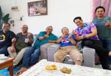 Seven men sit on a sofa in a bright living room, smiling and making finger-heart gestures; tea cups and snacks sit on the coffee table in front of them.