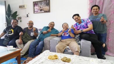 Seven men sit on a sofa in a bright living room, smiling and making finger-heart gestures; tea cups and snacks sit on the coffee table in front of them.