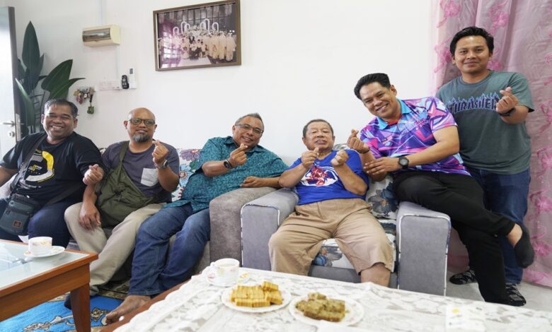 Seven men sit on a sofa in a bright living room, smiling and making finger-heart gestures; tea cups and snacks sit on the coffee table in front of them.