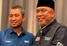 Two men smiling together at a indoor event, one in a blue shirt and the other in a dark jacket with a Malaysian flag patch and a black cap.