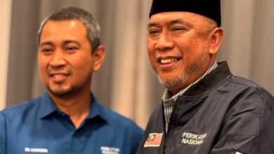 Two men smiling together at a indoor event, one in a blue shirt and the other in a dark jacket with a Malaysian flag patch and a black cap.