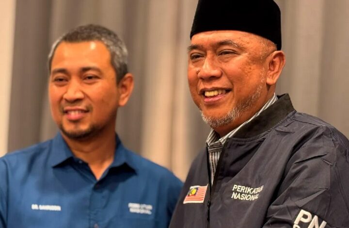 Two men smiling together at a indoor event, one in a blue shirt and the other in a dark jacket with a Malaysian flag patch and a black cap.