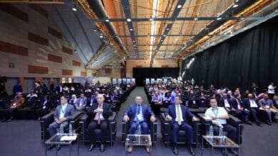 Conference hall with a large audience; five men in the front row wearing suits, water bottles on small tables.
