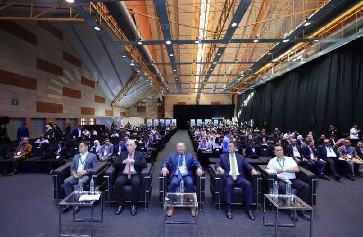 Conference hall with a large audience; five men in the front row wearing suits, water bottles on small tables.