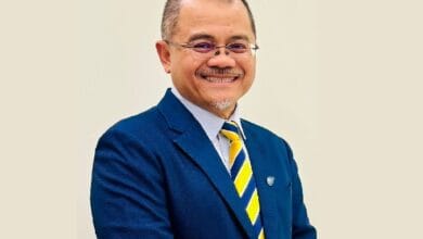 aimi zulhazmi - Wilayah.com.my Smiling man in a blue suit with a yellow and blue striped tie, standing against a light background.