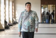 albert tei - Wilayah.com.my Man wearing a teal/gray patterned button-down shirt walks toward the camera in a sunlit hallway; luggage and blurred people in the background show an airport or hotel setting.