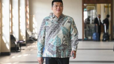 albert tei - Wilayah.com.my Man wearing a teal/gray patterned button-down shirt walks toward the camera in a sunlit hallway; luggage and blurred people in the background show an airport or hotel setting.