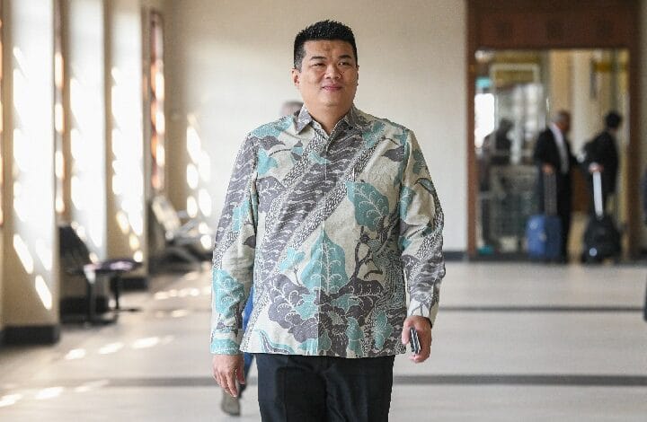 Man wearing a teal/gray patterned button-down shirt walks toward the camera in a sunlit hallway; luggage and blurred people in the background show an airport or hotel setting.