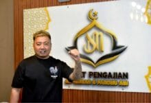 along - Wilayah.com.my Man with blond hair and tattooed left forearm wearing a black T-shirt, posing with his fist raised in front of a white wall sign featuring a gold crescent logo and text.