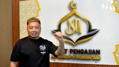 along - Wilayah.com.my Man with blond hair and tattooed left forearm wearing a black T-shirt, posing with his fist raised in front of a white wall sign featuring a gold crescent logo and text.
