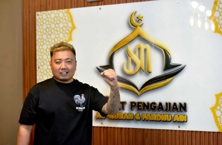 along - Wilayah.com.my Man with blond hair and tattooed left forearm wearing a black T-shirt, posing with his fist raised in front of a white wall sign featuring a gold crescent logo and text.
