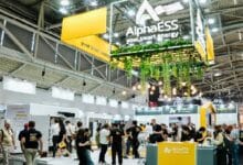 Trade show booth for AlphaESS energy with yellow branding, hanging plants, and attendees exploring displays at a busy expo hall.