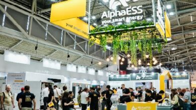 Trade show booth for AlphaESS energy with yellow branding, hanging plants, and attendees exploring displays at a busy expo hall.