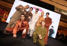 alpha mpop - Wilayah.com.my Group of eight male models posing on a fashion show runway, wearing bold, retro-colored suits with a bright screen backdrop behind them.