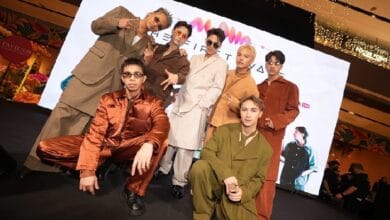 Group of eight male models posing on a fashion show runway, wearing bold, retro-colored suits with a bright screen backdrop behind them.
