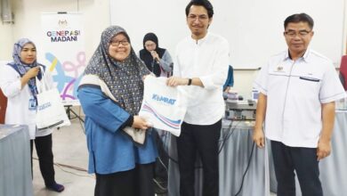 amk felda - Wilayah.com.my Man in a white shirt hands a branded bag to a smiling woman in a blue hijab at a community event; others look on.