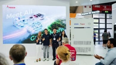 Four people pose for a photo in front of a large exhibition screen that reads 'Meet All' at a white trade-show booth; audience members sit nearby.