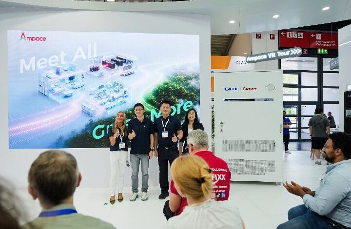 Four people pose for a photo in front of a large exhibition screen that reads 'Meet All' at a white trade-show booth; audience members sit nearby.