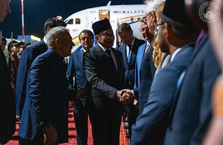 Officials in suits on a red carpet at night shake hands in front of a parked airplane, suggesting a formal arrival event.