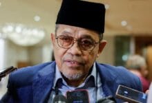anwar japan 2 - Wilayah.com.my Portrait of a middle-aged man wearing a black traditional cap and glasses, speaking to reporters with handheld microphones nearby.