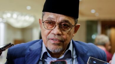anwar japan 2 - Wilayah.com.my Portrait of a middle-aged man wearing a black traditional cap and glasses, speaking to reporters with handheld microphones nearby.