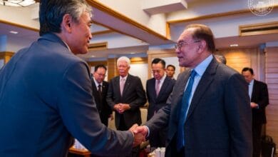 Two businessmen in suits shake hands in a formal meeting, with colleagues watching in a conference room.