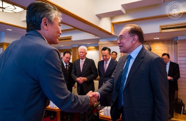 anwar japan 2 - Wilayah.com.my Two businessmen in suits shake hands in a formal meeting, with colleagues watching in a conference room.