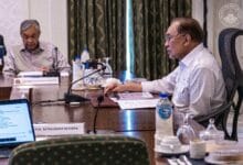 Two men in a formal meeting at a conference table with microphones, papers, and water bottles on the table.