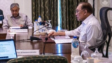 Two men in a formal meeting at a conference table with microphones, papers, and water bottles on the table.