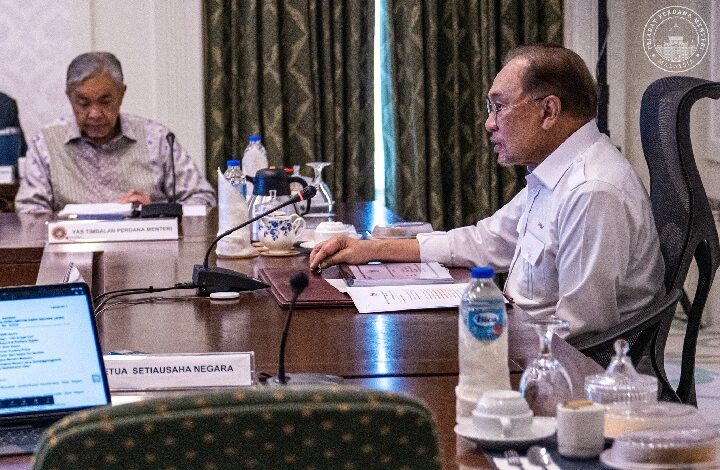Two men in a formal meeting at a conference table with microphones, papers, and water bottles on the table.