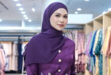 anzalna nasir - Wilayah.com.my Woman wearing a purple hijab and matching buttoned top, posing in a clothing store with colorful garments in the background
