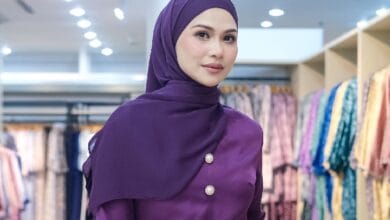 Woman wearing a purple hijab and matching buttoned top, posing in a clothing store with colorful garments in the background