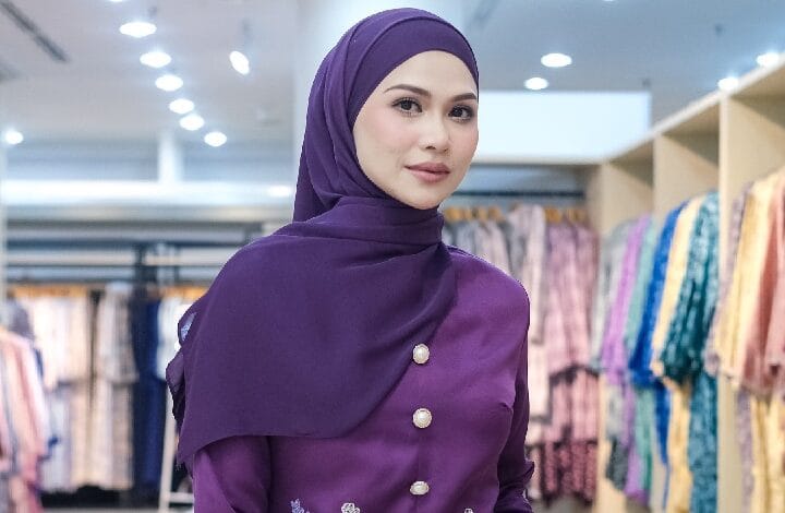 Woman wearing a purple hijab and matching buttoned top, posing in a clothing store with colorful garments in the background