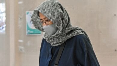 asnida abdul daim - Wilayah.com.my Woman wearing a patterned gray hijab and gray face mask, glasses, and a navy top, carrying a shoulder bag while walking in an indoor corridor.