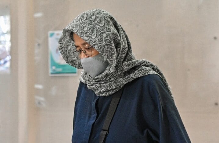 asnida abdul daim - Wilayah.com.my Woman wearing a patterned gray hijab and gray face mask, glasses, and a navy top, carrying a shoulder bag while walking in an indoor corridor.