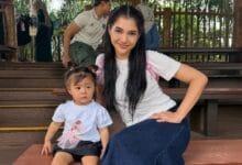 Young woman with long dark hair sits on wooden steps beside a toddler wearing a light blue shirt, posing for a photo outdoors on a shaded deck area.