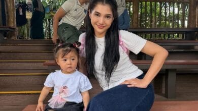 atikah suhaime - Wilayah.com.my Young woman with long dark hair sits on wooden steps beside a toddler wearing a light blue shirt, posing for a photo outdoors on a shaded deck area.