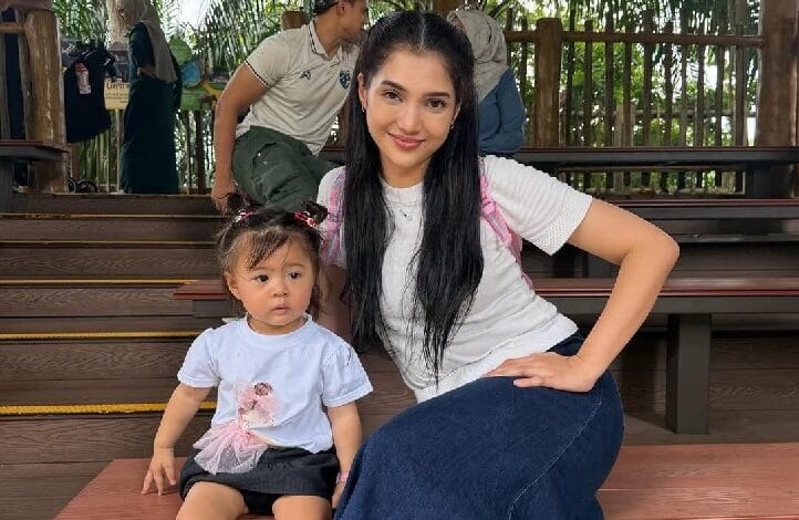 Young woman with long dark hair sits on wooden steps beside a toddler wearing a light blue shirt, posing for a photo outdoors on a shaded deck area.
