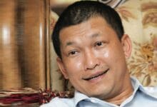 azlan johar - Wilayah.com.my Man with short black hair wearing a light blue button-down shirt, smiling while seated on a patterned sofa in a cozy room