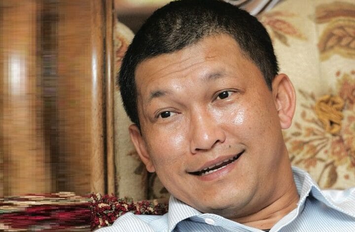 azlan johar - Wilayah.com.my Man with short black hair wearing a light blue button-down shirt, smiling while seated on a patterned sofa in a cozy room