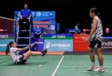badminton malaysia - Wilayah.com.my Badminton match scene: a player on the floor reaching with her racket toward the standing opponent while an umpire sits on a high chair nearby. Sponsors visible in background.