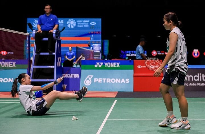 badminton malaysia - Wilayah.com.my Badminton match scene: a player on the floor reaching with her racket toward the standing opponent while an umpire sits on a high chair nearby. Sponsors visible in background.