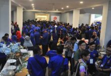 bn barisan nasional - Wilayah.com.my Crowd of people in blue shirts gathered in a large indoor event space, with tables and displays along the sides.