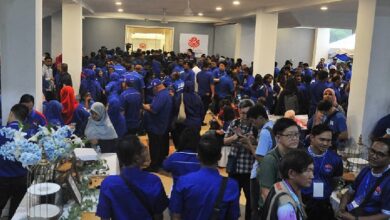 bn barisan nasional - Wilayah.com.my Crowd of people in blue shirts gathered in a large indoor event space, with tables and displays along the sides.