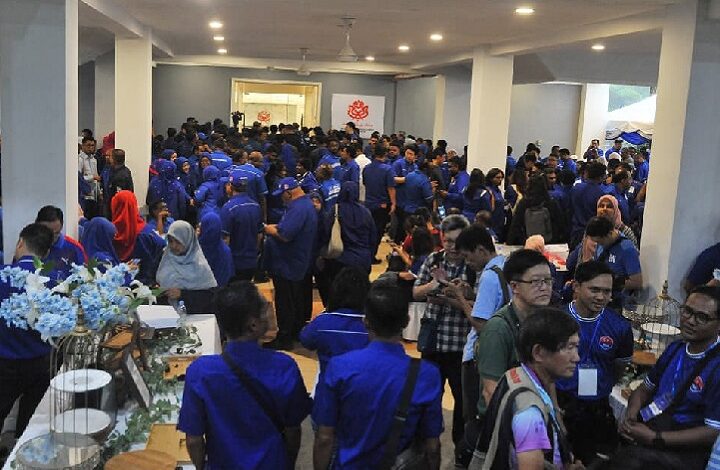 Crowd of people in blue shirts gathered in a large indoor event space, with tables and displays along the sides.