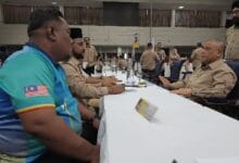 bn perak - Wilayah.com.my Men in tan uniforms sit at a long conference-table in a large hall, foregrounded by a man in a turquoise shirt with a Malaysia flag patch on the sleeve.