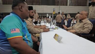 Men in tan uniforms sit at a long conference-table in a large hall, foregrounded by a man in a turquoise shirt with a Malaysia flag patch on the sleeve.