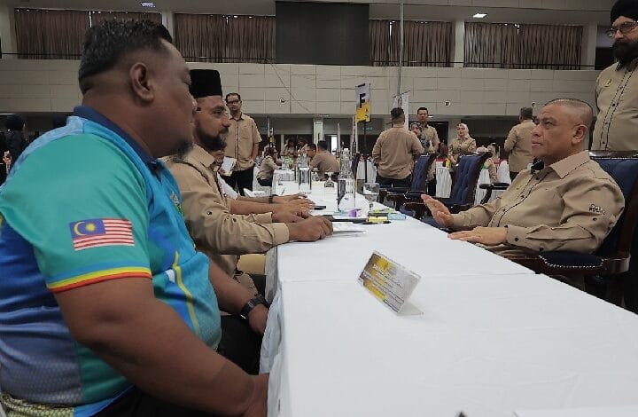 Men in tan uniforms sit at a long conference-table in a large hall, foregrounded by a man in a turquoise shirt with a Malaysia flag patch on the sleeve.