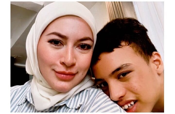 Close-up of a woman in a white hijab smiling with a young man leaning in for a selfie beside her.
